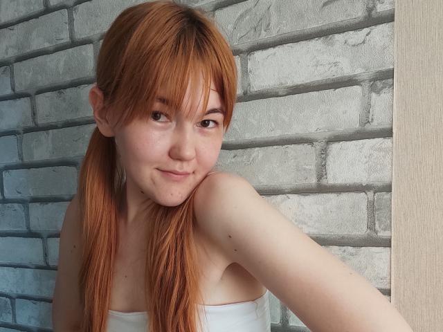 SheritaChieng with red pigtails, smiling, wearing a white strapless top, showing her small tits and freckles, leaning against a brick wall.