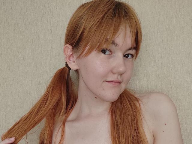 SheritaChieng, met rode vlechtjes, lichte huid, kleine borsten, sproeten en een lichte glimlach, houdt haar haar vast, voor een beige muur. Haar bruine ogen kijken schuin..