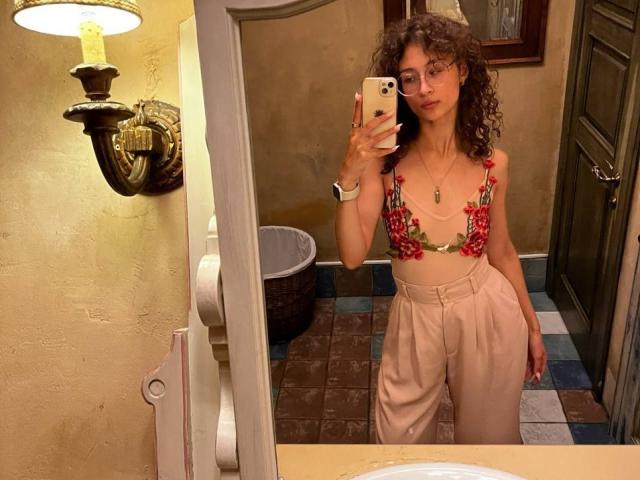 AlyceErway with curly brown hair, glasses, and a red floral bikini top, showing her tits, wearing high-waisted beige pants, taking a mirror selfie in a bathroom.