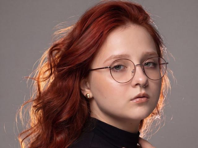 SaraMunewe heeft rood haar, ronde brillen, een neusring en een zwarte coltrui. Ze heeft een serieuze uitdrukking, een lichte huid en een kleine gouden oorbelle in haar linker oor..