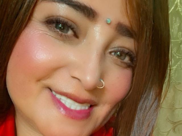 AlessadraJoness hat ein hübsches Gesicht mit braunen Augen, einem Nasenring, einem türkisblauen Punkt auf der Stirn und pinkem Lippenstift, lächelt mit rosigen Wangen und hat glattes, braunes Haar..