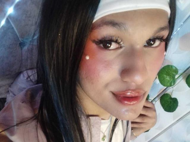 PamelaXFox hat glänzende, rosafarbene Lippen, schwarzen Eyeliner und einen weißen Haarreif, sowie schwarzes Haar, gerötete Wangen und kleine, weiße Punkte auf der Nase. Sie trägt eine rosa Kapuzenjacke..