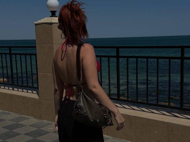 AlexXas with red hair, back to the camera, in a red halter top, black skirt, and brown bag, standing by a seafront railing, showing her bare ass.