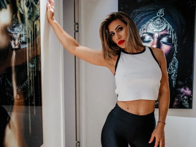 SweetBelle, con addominali scolpiti, indossa un top corto bianco senza maniche e leggings neri, ha il rossetto rosso e un'acconciatura con frangia laterale, ed è in piedi con un braccio appoggiato a un muro..
