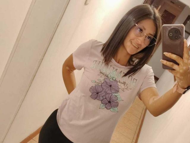 SharissWilliam, con i capelli castano corti, occhiali, che sorride, indossa una maglietta rosa con fiori e pantaloncini neri, sta scattando un selfie allo specchio con un telefono rosa in un corridoio..
