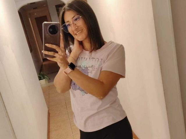 SharissWilliam mit Brille, lächelnd, hält ein Telefon, trägt ein weißes T-Shirt, schwarze Hosen und eine pinke Uhr, macht ein Selfie im Spiegel und zeigt ihr Gesicht und ihre Brust..