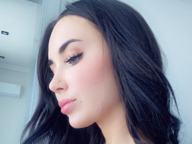 RinaBrown heeft zwart haar, dikke wenkbrauwen, lange wimpers, een roze blos en een klein beauty markje op haar bovenlip. Haar lippen zijn lichtjes open. Ze kijkt naar beneden..