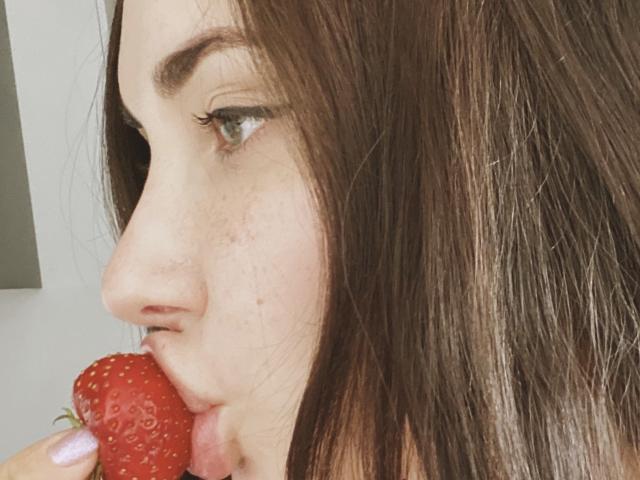 RinaBrown, met bruin haar, groene ogen en sproeten, eet een rode aardbei met haar mond. Haar lippen zitten om de onderkant van de aardbei. Haar neus is iets omhoog gekeerd..