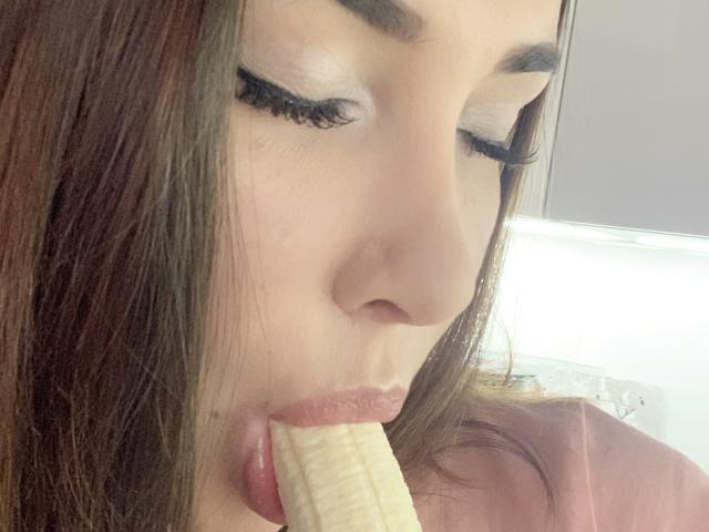 Het gezicht van RinaBrown, met bruin haar, witte make-up en lange wimpers, terwijl ze een banaan eet, waarbij haar roze lippen eromheen zijn gevouwen..