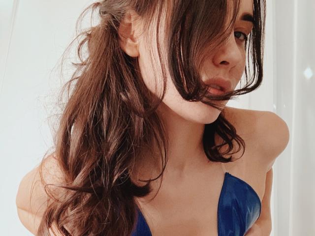 RinaBrown met golvend bruin haar, een blauwe bikini-top, volle lippen en een licht gekanteld hoofd, die haar borsten laat zien. Zacht licht op haar gezicht, ze ziet er sexy uit..