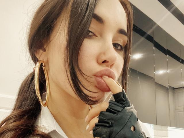 RinaBrown met volgehouden lippen, bruin haar in een paardenstaart, gouden oorringen, een zwarte handschoen zonder vingers, waarbij ze haar tong tegen haar vinger houdt, een wit shirt, lichte huid, knippend met haar oog..