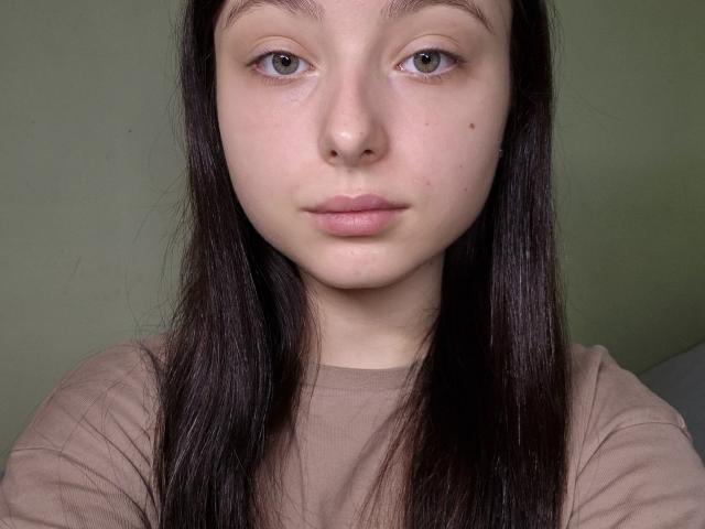 Il viso di ManaPlachecki, pelle pallida, capelli neri lisci, occhi verde chiaro, labbra rosa delicatamente colorate, naso piccolo, qualche piccolo segno di acne, indossa una camicia beige, espressione neutra..