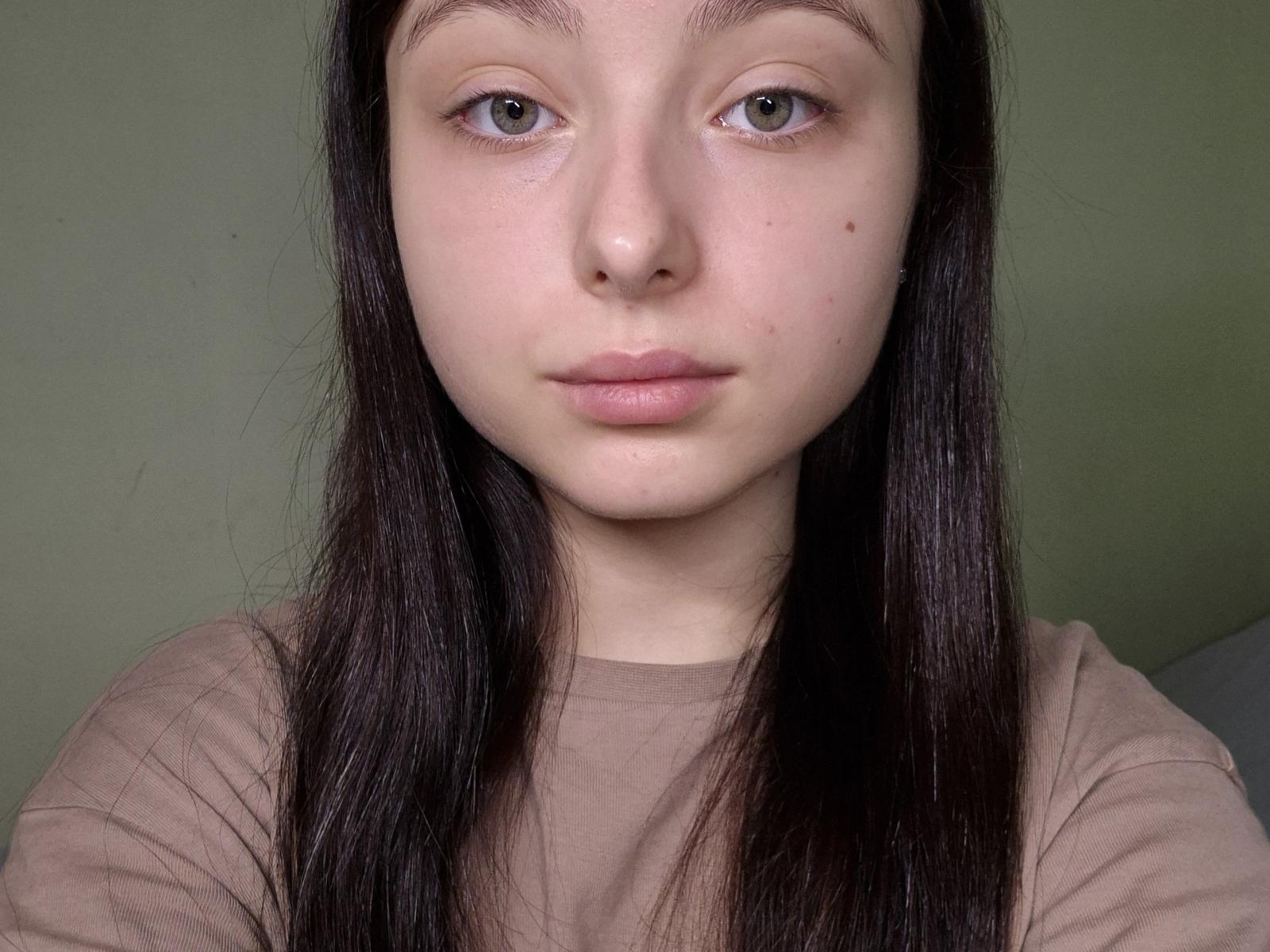 O rosto de ManaPlachecki, pele clara, cabelo castanho escuro e liso, olhos verde claro, lábios rosados delicados, nariz pequeno, algumas pequenas manchas de acne, vestindo uma camisa bege, expressão neutra..