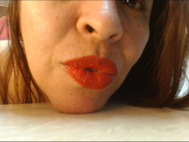 LizethBells' Gesicht liegt auf einem Tisch, ihre roten, geschwollenen Lippen sind leicht geöffnet und ihre Nase zeigt nach oben. Ihr braunes Haar ist teilweise sichtbar..