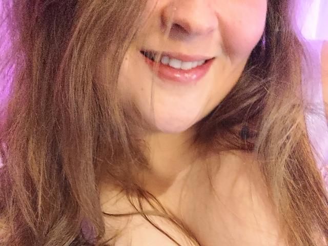 Lilysweetxx a les cheveux châtains clairs, un sourire discret avec des lèvres roses brillantes, et une décolleté visible. Sa peau est claire avec une légère teinte rosée. Ses seins sont partiellement visibles..