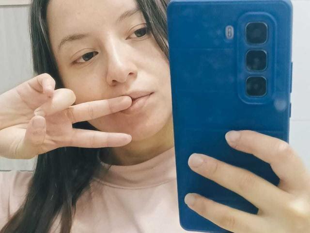 VioletaNova with long dark hair, pink shirt, holding blue phone, making a peace sign with finger to her lips, showing a subtle smile, looking into the camera.