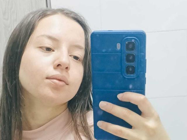 VioletaNova with long brown hair, holding a blue phone, pouting slightly, wearing a pink shirt, in a bathroom selfie. Her hand covers half the phone.