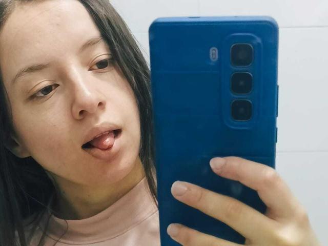 VioletaNova's face with long brown hair, pale skin, and tongue slightly out, holding a blue phone with three cameras, wearing a pink shirt, in a selfie.