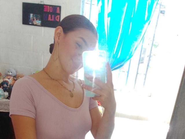 AbbyMilers with brown hair in a pink top, holding a phone in front of her face, smiling, showing her small tits, wearing gold earrings and a necklace.