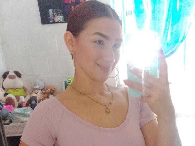 AbbyMilers with brown hair, wearing a pink top, smiling at a mirror selfie, holding a phone, gold necklace, hoop earrings, soft light, bright blue curtain in background.
