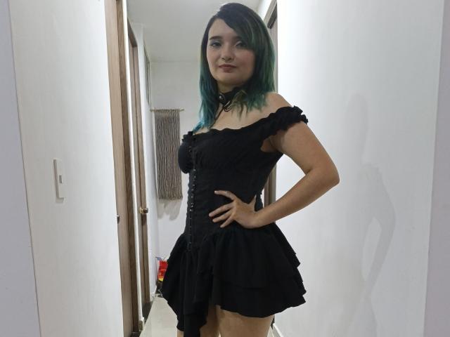 NaommyCarter with blue-green hair, black off-shoulder dress, hand on hip, small tits, black choker, standing in hallway, smiling, fair skin, slender, cute, confident.