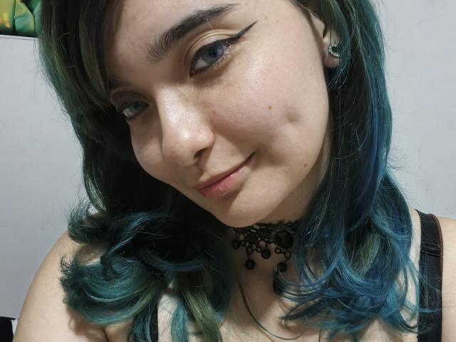 NaommyCarter has teal-blue hair, black eyeliner, and a slight smile. She's wearing a black lace choker and a strap showing. Her tits are partially visible.