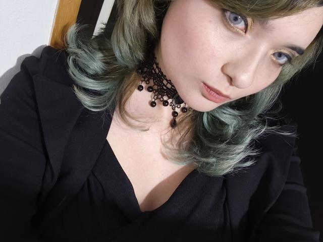 NaommyCarter with greenish-blue hair, blue eyes, and a black choker with beads, wearing a black top, showing a small cleavage, and a subtle pout.
