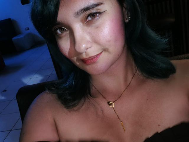 NaommyCarter has teal hair, pink cheeks, and a slight smile. She's wearing a necklace with a small square and a thin gold chain, showing her tits.
