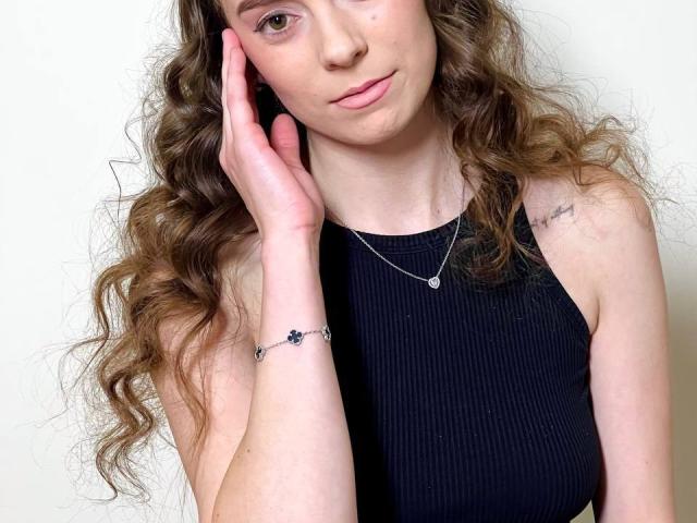 NicoleInfinity, avec ses cheveux bruns bouclés, portant un débardeur noir côtelé, touchant sa joue avec sa main gauche, un collier en argent et un petit tatouage sur son épaule droite..