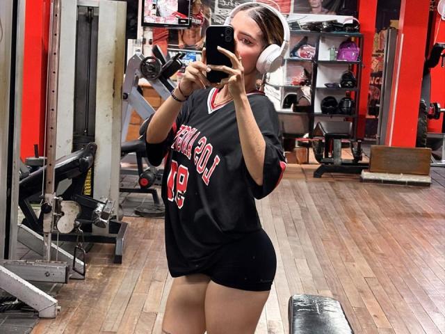 SecretLoversVIP dans une salle de sport, portant un maillot noir "New York", des écouteurs blancs, sans pantalon, prenant un selfie devant un miroir, montrant des fesses galbées et un espace entre les cuisses, sol en bois..