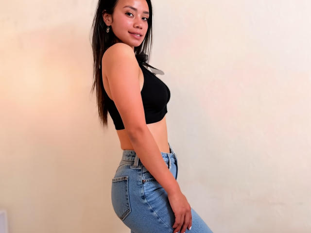 AlinaVale mit langen, schwarzen Haaren, die ein schwarzes Crop-Top und blaue Jeans trägt, wobei ihr Hintern und ihre kleinen Brüste sichtbar sind, lächelt leicht und steht seitlich zu einer schlichten Wand..