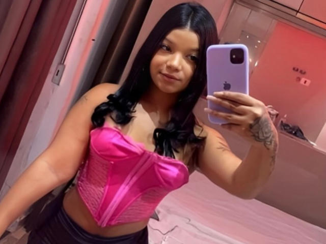 Tattuada69, com cabelo preto, corpete rosa e tatuagem no braço direito, tirando uma selfie com um iPhone, mostrando os seios e o bumbum pequeno. Quarto com luzes rosa e vermelhas..