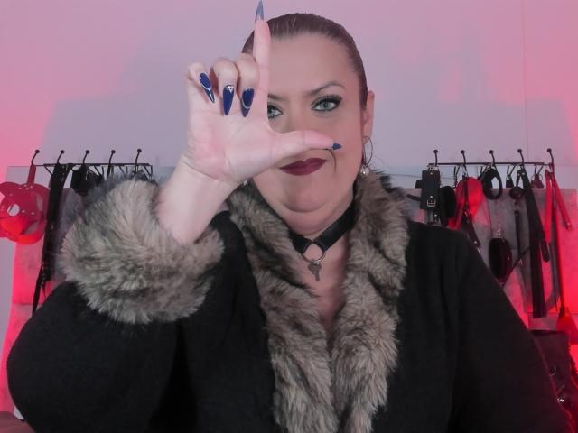 VanesaMoka, avec du rouge à lèvres foncé, des ongles bleus et une veste noire bordée de fourrure, faisant un geste de la main, portant un collier noir, sur un fond rose éclairé avec du matériel BDSM..