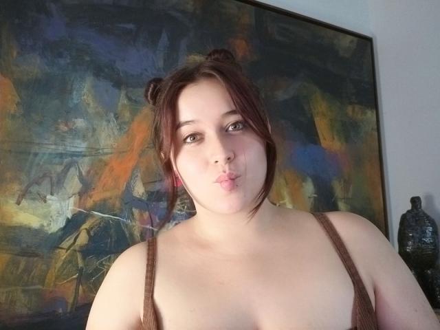 AngelaMonticelli with red hair in double buns, puffy lips, brown tank top, showing her boobs, standing in front of a colorful abstract painting, holding a water bottle.