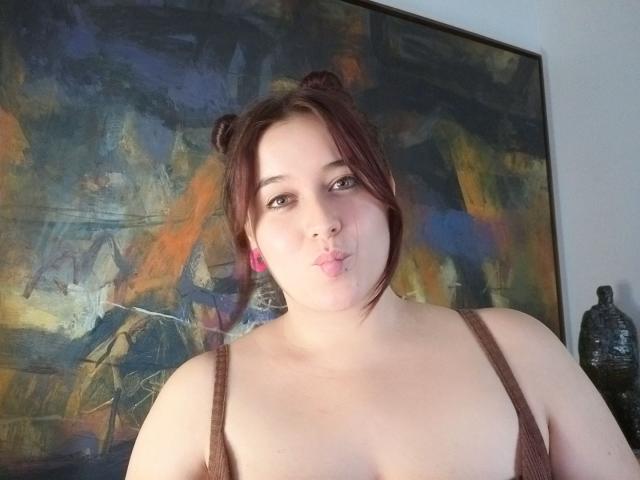 Angela Monticelli, avec ses cheveux bruns bouclés attachés en chignons, un débardeur marron laissant entrevoir ses seins, des boucles d'oreilles roses, et affichant une moue boudeuse, dans un cadre intérieur décontracté..