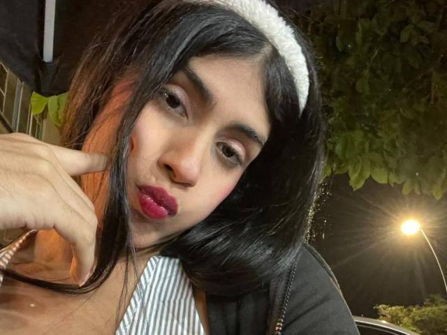 ValeryTranss mit dunklem Haar, weißem Stirnband, pinkem Lippenstift und hochgezogenen Mundwinkeln, die mit dem Finger auf ihre Wange zeigt, trägt ein gestreiftes Hemd und eine schwarze Jacke, im Hintergrund ist eine nächtliche Straßenlaterne zu sehen..
