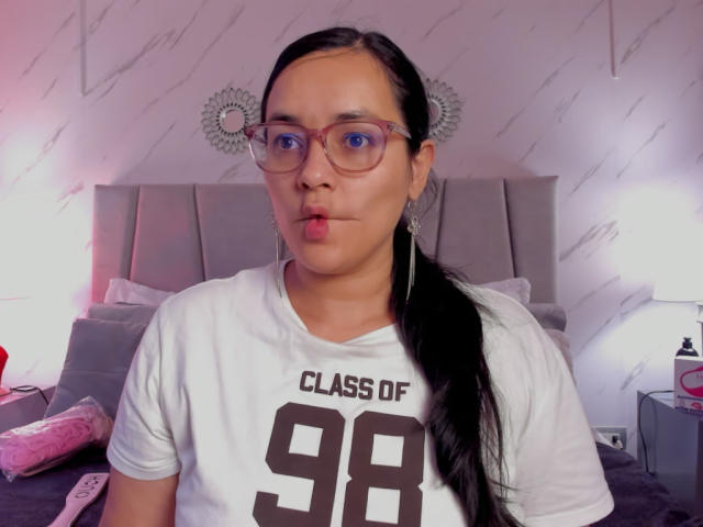 MichellMonet, lèvres pulpeuses, portant des lunettes, chemise blanche avec le numéro 98, queue de cheval noire, lit avec une tête de lit grise, jouet rose à gauche, lampe blanche à droite..
