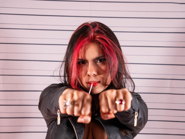 MaraCortes with red-black hair, black jacket, pointing with finger, red lipstick, rings, and zipped jacket, showing off her tits, looking intense, with a cigarette in her mouth.