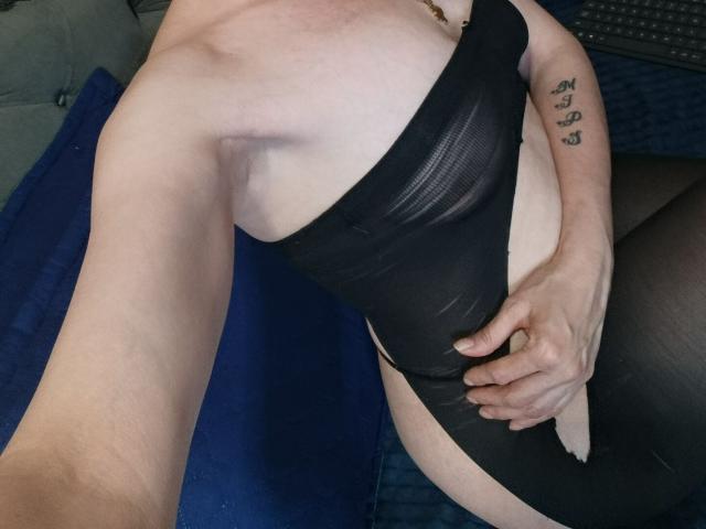 RubiAnderson wearing a black sheer one-piece, showing her tits and ass, with one hand on her crotch, tattoo on her right arm, sitting on a blue surface.
