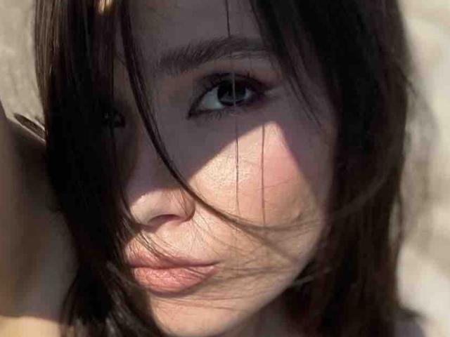 El rostro de SelenaAllure, con cabello castaño oscuro que cubre parcialmente su ojo azul, labios rosados suaves y piel clara. La luz del sol proyecta sombras sobre su mejilla. Parece pensativa y atractiva..