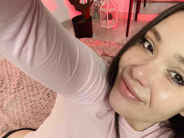 SelenaAllure, con una sonrisa, camisa rosa de manga larga y cabello oscuro, sosteniendo un brazo para tomarse un selfie, alfombra rosa, luz roja y un farol blanco en el fondo..