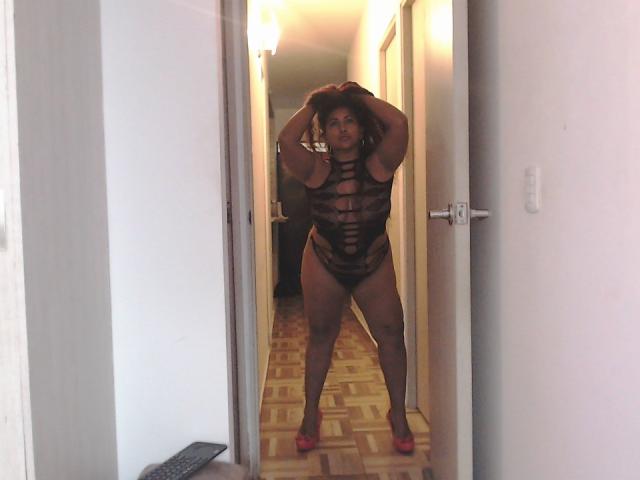 YiyaHot69 è in un corridoio, indossa un set di lingerie nero con cinturini e tacchi rosa, ha le mani tra i capelli ricci, mostra un sedere grande e cosce formose, e sorride alla telecamera..