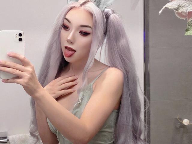 Icyvai-hot, com cabelos longos, prateado-rosados em maria-chiquinhas, com a língua de fora, segurando um telefone, vestindo uma blusa verde, com a mão no peito, em um banheiro..