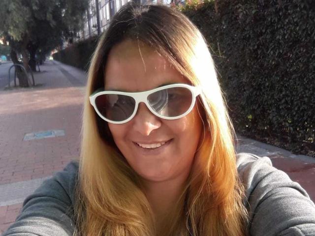 DubraskaBella hat langes, blondes Haar, eine Brille mit weißem Rahmen und ein Lächeln. Sie trägt ein graues Hemd und steht im Freien in der Sonne. Ihre Brüste sind nicht sichtbar. Sie lächelt direkt in die Kamera..