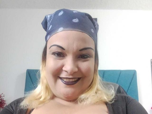 DubraskaBella has a chubby face, dark purple lipstick, blonde hair, blue bandana with white patterns, and is smiling. She's wearing a black top, and has visible dark eyebrows.