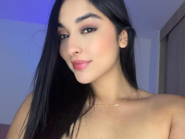 Emma Waltter hat langes, schwarzes Haar, braune Augen und rosafarbene Lippen mit einem leichten Lächeln. Sie ist oben ohne, trägt eine goldene Halskette und hat einen hellen Hautton..