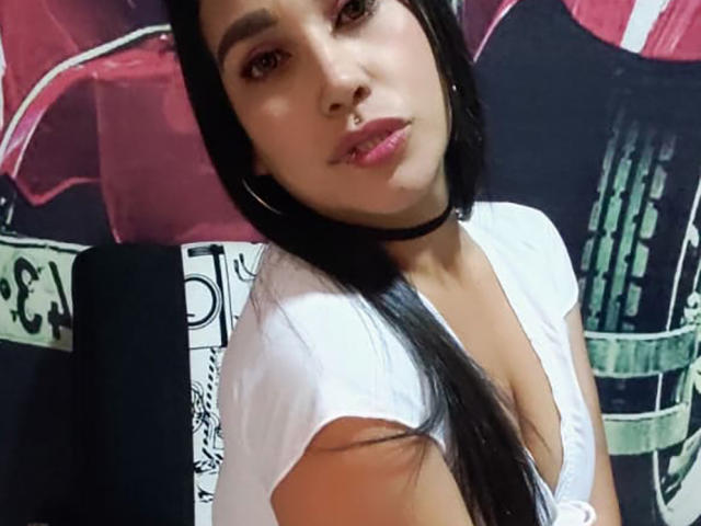 EdenIvyLm, con cabello largo y negro, vestida con una blusa blanca de cuello en V pronunciado que muestra sus senos, un collar negro y aros grandes, mirando a la cámara con un ligero puchero..