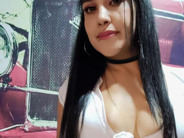 EdenIvyLm, com longos cabelos pretos, vestindo uma blusa branca de decote baixo que mostra seus seios grandes, um colar preto e batom rosa, olhando para a câmera com um leve sorriso..