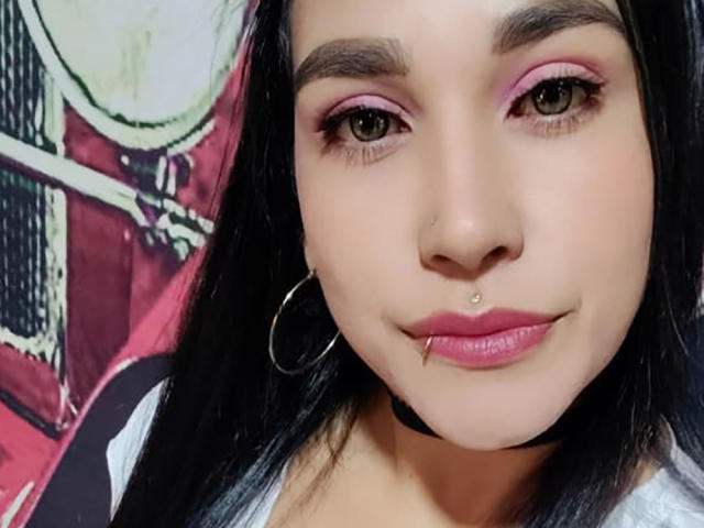 EdenIvyLm tem sombra rosa nos olhos, um piercing no nariz, um piercing no lábio e usa um colar preto com brincos de argola. Seu cabelo escuro contrasta com sua maquiagem rosa..