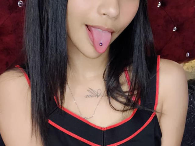 YayaLm with black hair, tongue out, pink tongue piercing, black and red dress, chest tattoo, and a necklace, showing her ass and tits.